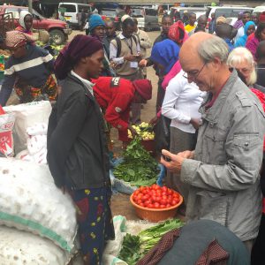Man with smartphone in crowded market, with woman selling fresh produce and bags of dried goods in Tanzania