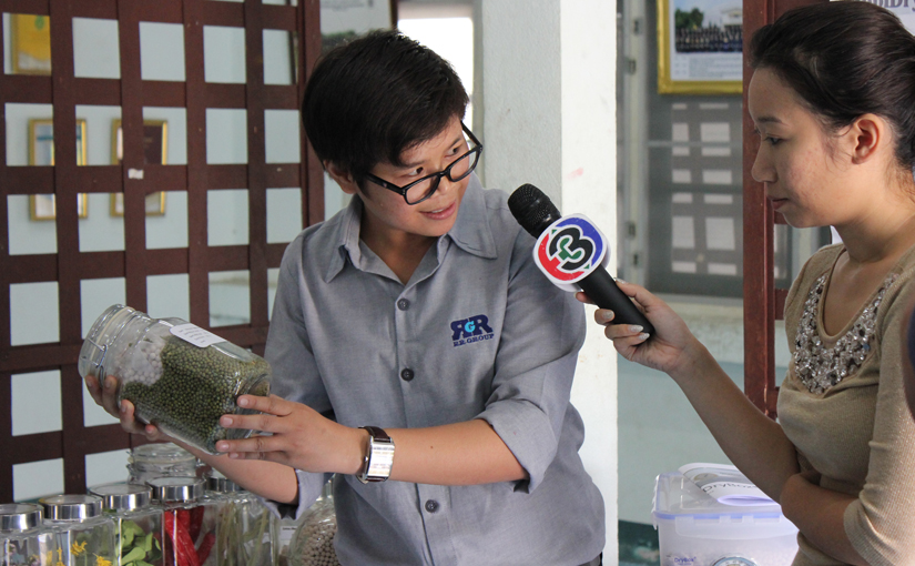 Representative shows jar of seeds and drying beads to television reporter in Thailand.