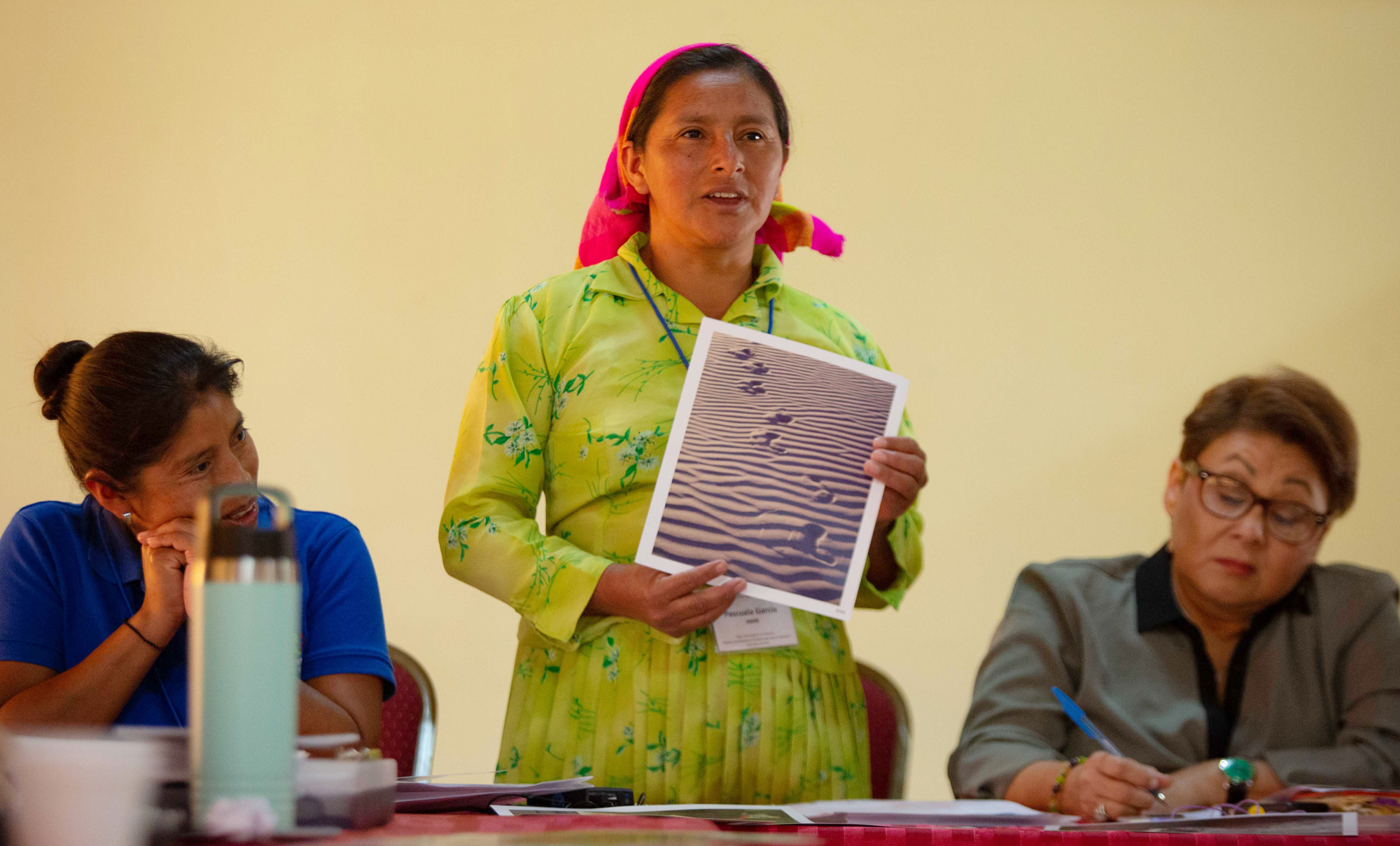 Woman in traditional dress stands at a meeting to speak, holding a photo, with professional women seated on either side of her