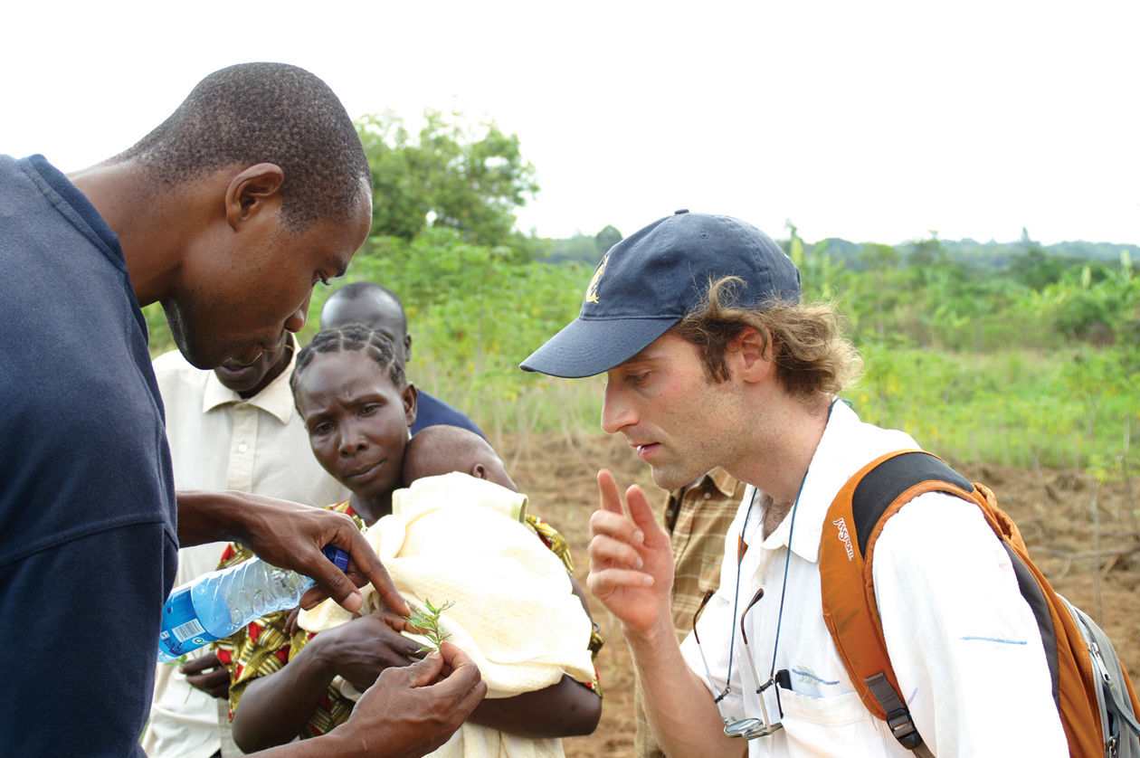 Kenyan staff member shows American student plant detail with farmers in background, in Kenya
