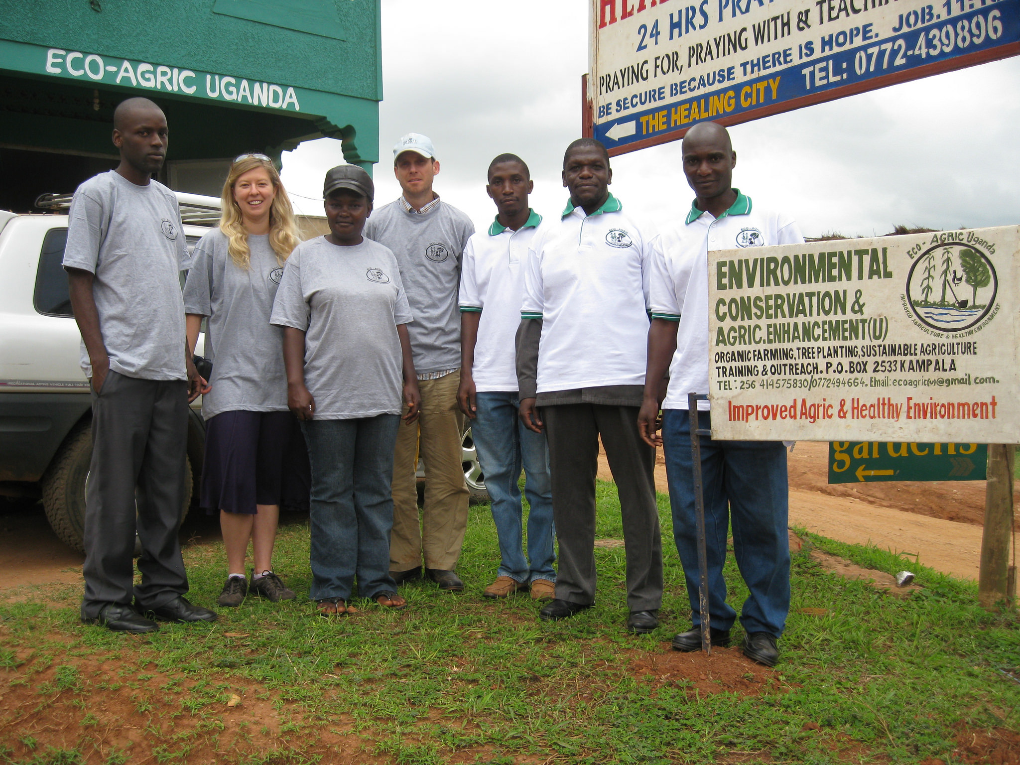 Group of Ugandan staff and volunteers, Shapland, and Peach-Fine in matching shirts stand next to a sign that reads "environmental conservation & agric.enhancement (U)...improved agric & healthy environment"