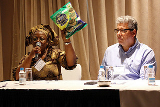 Woman sitting behind table with microphone holds up a bag, man sits next to her