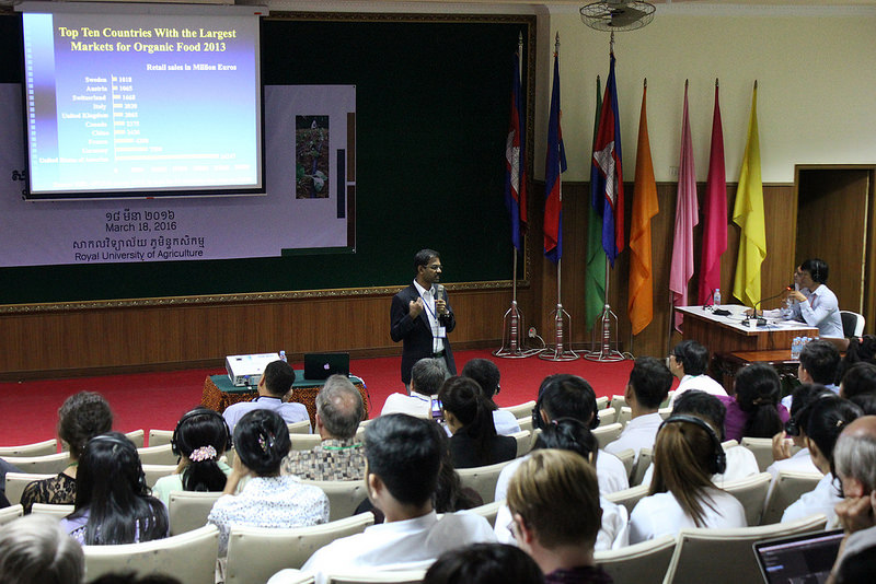 Govindasamy stands in front of a seated crowd, holding a microphone, in a lecture hall. Screen reads "top ten countries with the largest markets for organic food 2013," seven flags in the right of the room
