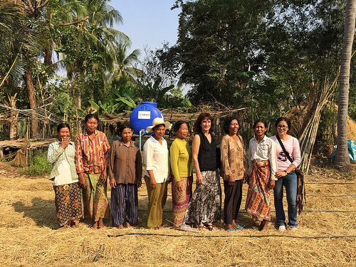 Women stand in a field in a line, drip tape at their feet, behind them a water tank