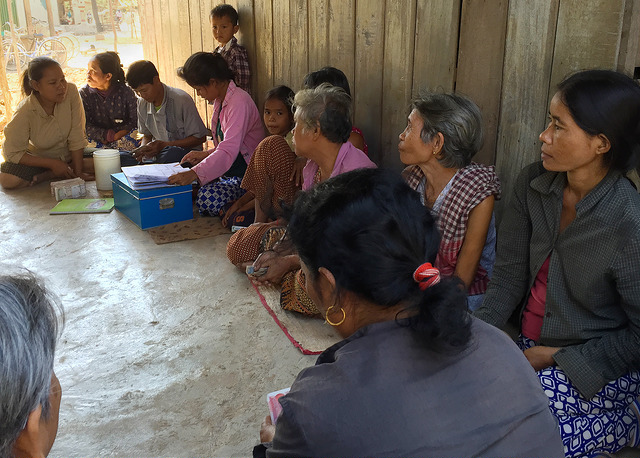 Women sit and look on as one woman peruses paper documents