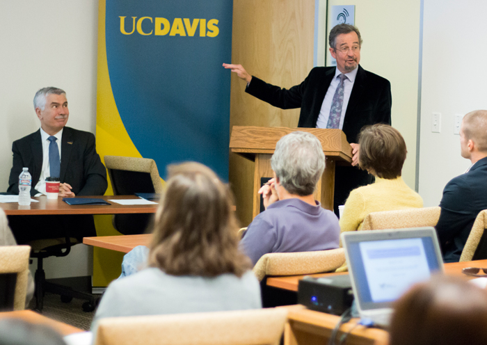 Man gesturing at podium, audience members and panelist look on