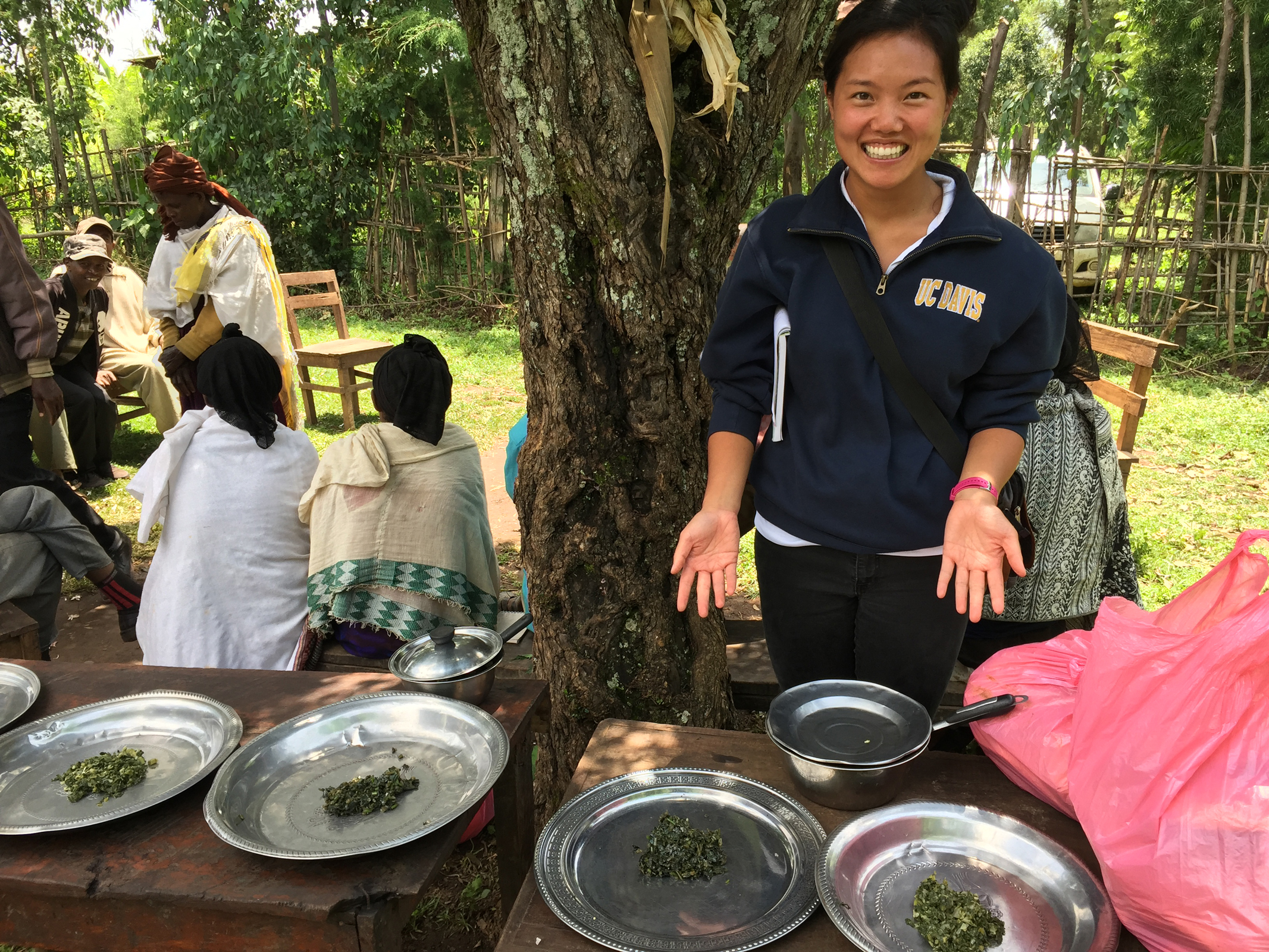 UC Davis student gestures to plates of cooked leafy greens, outside with farmers in Ethiopia