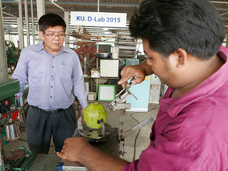 In front of a sign that reads "D-Lab", man uses machine to open a coconut, other man looks on