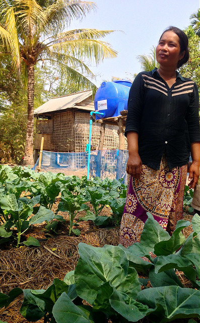 woman farmer stands in field among leafy vegetables and mulch, with water tank in background