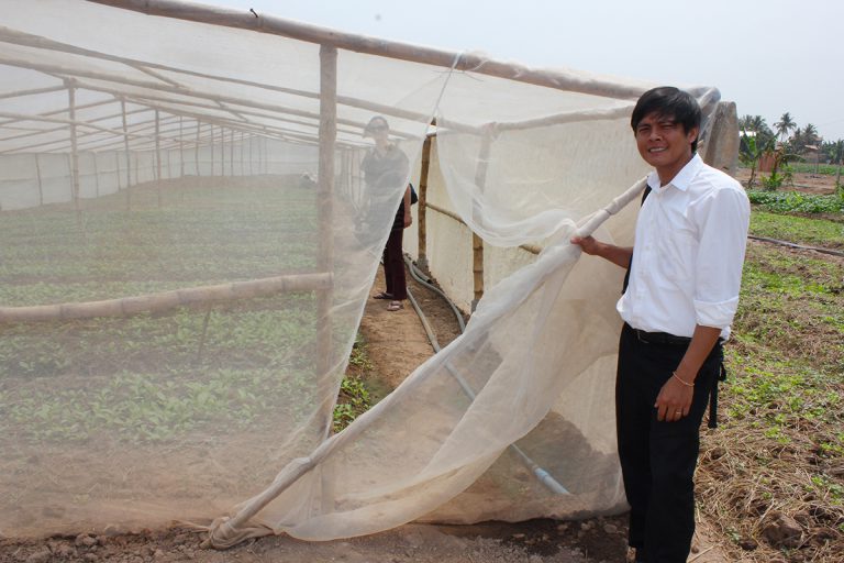 In a vegetable field, Thort holds open the net door of a net house, with someone standing inside among the vegetable rows