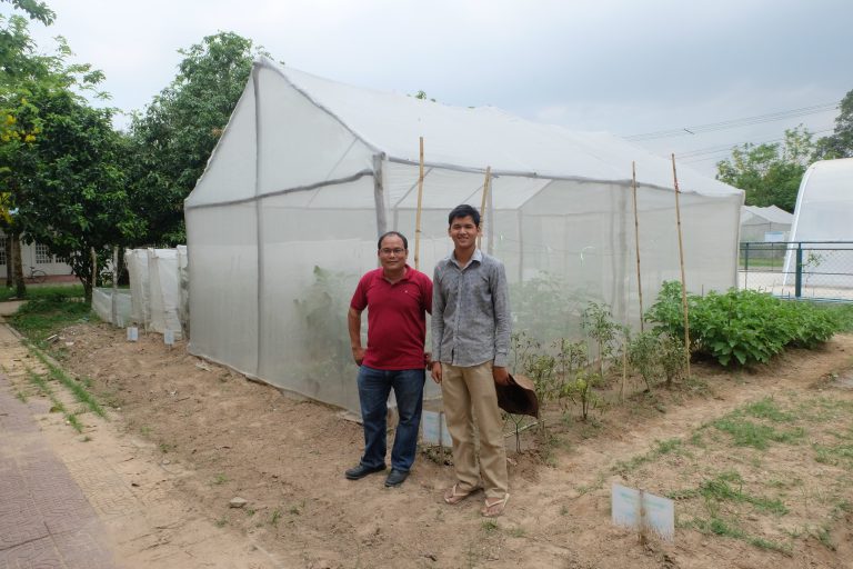 Two university scientists stand in front of net structures, with vegetable plants growing inside.