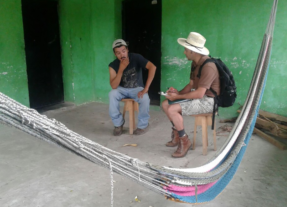 Young man wearing a backpack interviews an older man, in rural Honduras