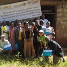 smiling group of farmers and researchers outdoors, in front of school house with usaid logo banner